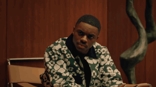 A Man In A Green Jacket Sits In A Chair In Front Of A Wooden Wall GIF
