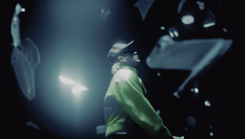 A Man In A Green Jacket Stands In A Dark Room Looking Up GIF