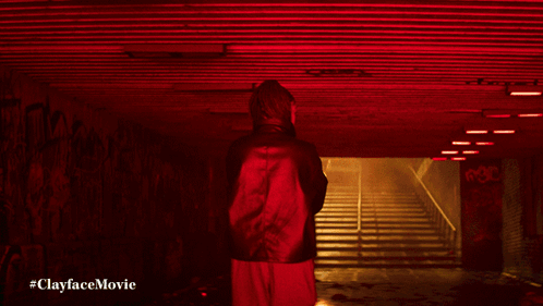 A Man In A Green Jacket Stands In A Dark Tunnel With The Words #Clayfacemovie Below Him GIF