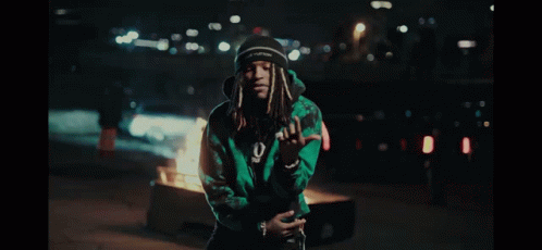 A Man In A Green Jacket Stands In Front Of A Burning Trash Can GIF