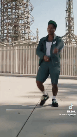 A Man In A Green Jumpsuit Is Dancing On A Sidewalk GIF