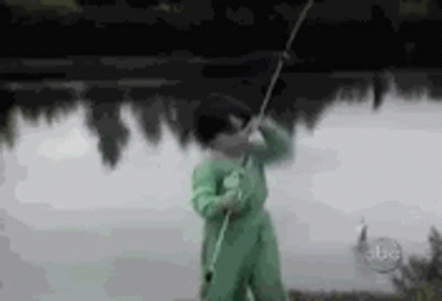A Man In A Green Jumpsuit Is Holding A Fishing Rod In Front Of A Lake . GIF