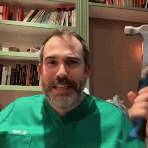 A Man In A Green Scrub Top Holds A Hammer In Front Of A Bookshelf GIF