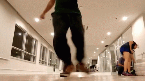 A Man In A Green Shirt And Black Pants Is Walking In A Hallway GIF