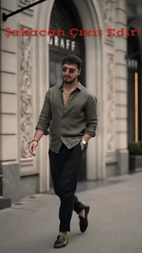 A Man In A Green Shirt And Black Pants Walks Down A Sidewalk In Front Of A Building That Says Graff GIF