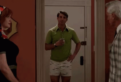 A Man In A Green Shirt And White Shorts Is Standing In Front Of A Door Holding A Cup Of Coffee . GIF