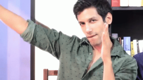 A Man In A Green Shirt Has His Arm Outstretched In Front Of A Bookshelf GIF