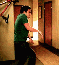 A Man In A Green Shirt Is Dancing In A Hallway With A Bike Hanging On The Wall Behind Him . GIF