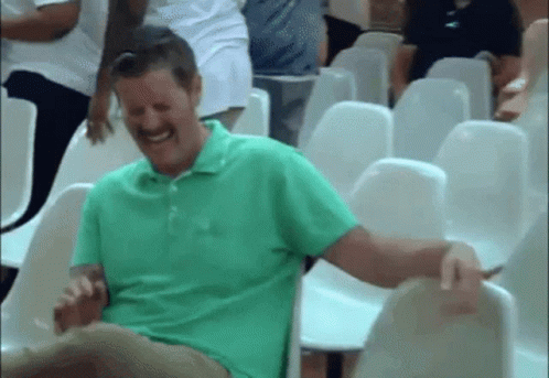 A Man In A Green Shirt Is Laughing While Sitting In A Row Of Chairs . GIF