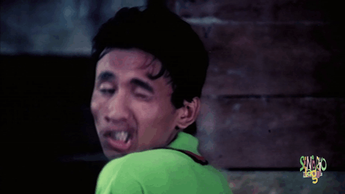 A Man In A Green Shirt Is Making A Funny Face In Front Of A Sign That Says ' Sword League ' GIF