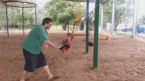 A Man In A Green Shirt Is Pushing A Child On A Swing At A Playground GIF