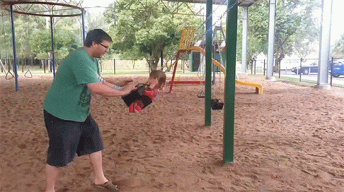A Man In A Green Shirt Is Pushing A Little Girl On A Swing GIF
