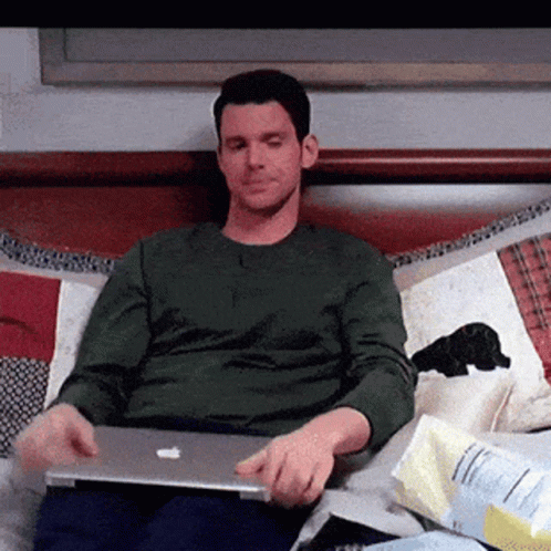 A Man In A Green Shirt Is Sitting On A Bed With A Laptop GIF