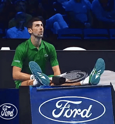A Man In A Green Shirt Is Sitting On A Ford Sign Holding A Tennis Racquet GIF