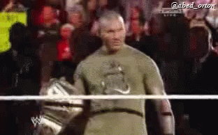 A Man In A Green Shirt Is Standing In A Wrestling Ring With A Crowd Behind Him . GIF