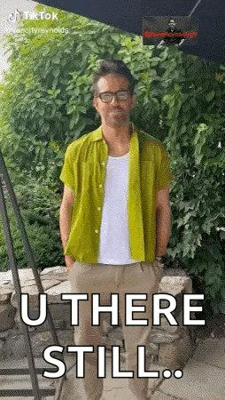 a man in a green shirt is standing in front of a stone wall with his hands in his pockets .