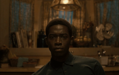 A Man In A Green Shirt Looks At The Camera In A Dark Room GIF