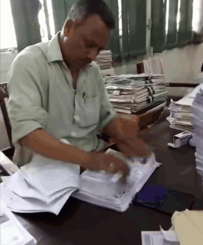 A Man In A Green Shirt Sits At A Desk With A Stack Of Papers GIF