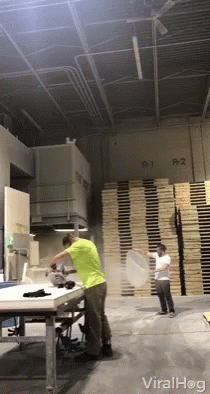 A Man In A Green Shirt Throws A Pizza In A Warehouse With A Stack Of Wooden Pallets In The Background GIF