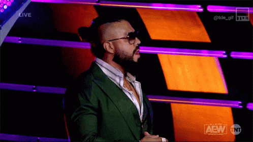 A Man In A Green Suit And Sunglasses Stands In Front Of A Purple And Orange Background That Says Aew On It GIF