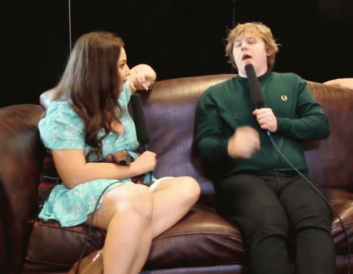 A Man In A Green Sweater Holds A Microphone While Sitting Next To A Woman GIF
