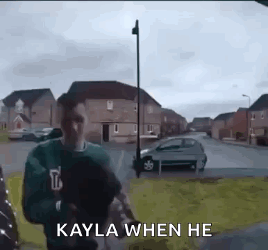 A Man In A Green Sweater Is Walking Down A Street With The Words `` Kayla When He '' Written On It . GIF