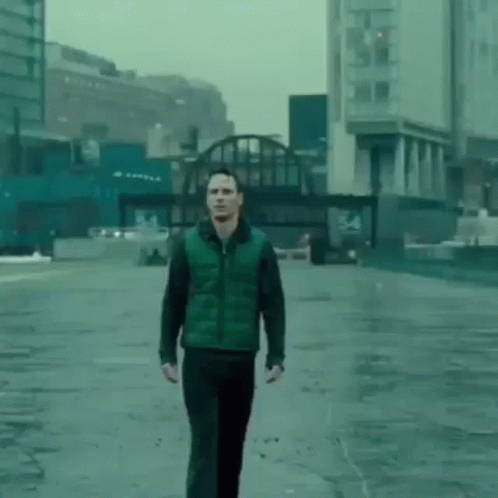 A Man In A Green Vest And Black Pants Is Walking Down A Wet Street In A City . GIF
