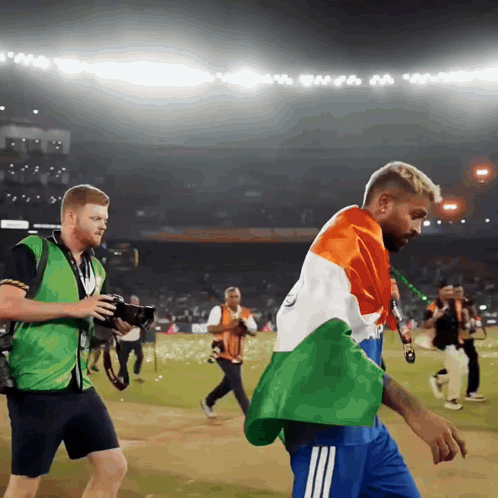 A Man In A Green Vest Is Taking A Picture Of Another Man In A Blue Shirt GIF