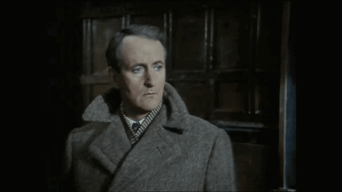 A Man In A Grey Coat Is Standing In Front Of A Door GIF