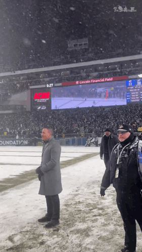 A Man In A Grey Coat Stands On A Snowy Field In Front Of A Scoreboard That Says Lincoln Financial Field GIF