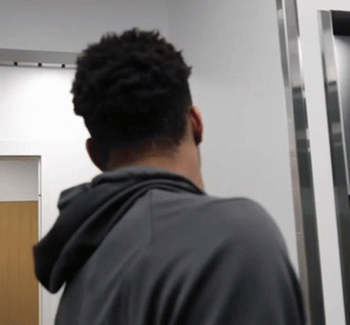 A Man In A Grey Hoodie Stands In A Hallway GIF