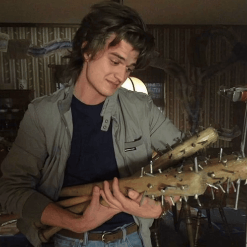 A Man In A Grey Jacket Is Holding A Bunch Of Sticks With Nails Sticking Out Of Them GIF