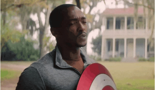 A Man In A Grey Shirt Is Holding A Red And White Shield In Front Of A House . GIF
