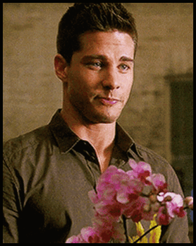 A Man In A Grey Shirt Is Holding Pink Flowers In His Hand GIF
