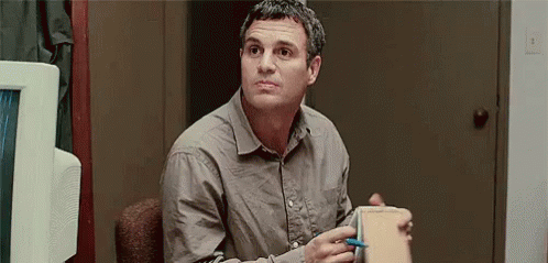 A Man In A Grey Shirt Is Sitting In A Chair Holding A Box And A Pen . GIF