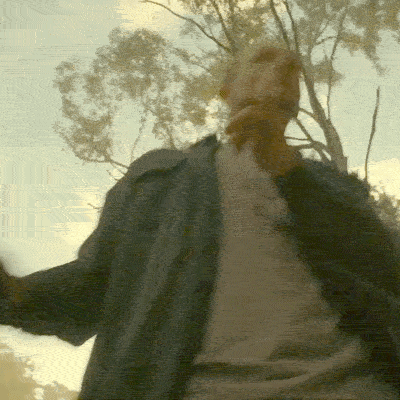 A Man In A Grey Shirt Is Standing In Front Of A Tree GIF