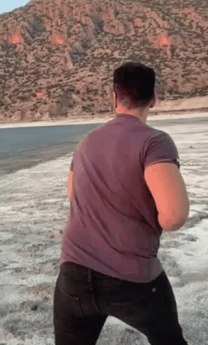A Man In A Grey Shirt Is Standing On A Beach With Mountains In The Background GIF
