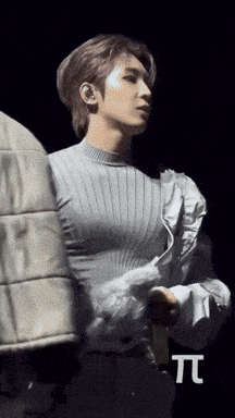 A Man In A Grey Sweater Is Standing In Front Of A Black Background With The Letter T. GIF