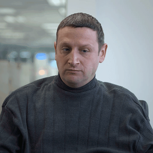 A Man In A Grey Sweater Looks At The Camera With A Serious Look On His Face GIF