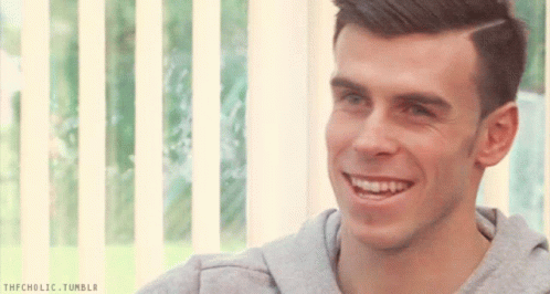 A Man In A Grey Sweatshirt Is Smiling In Front Of A Window With A Striped Blind Behind Him GIF