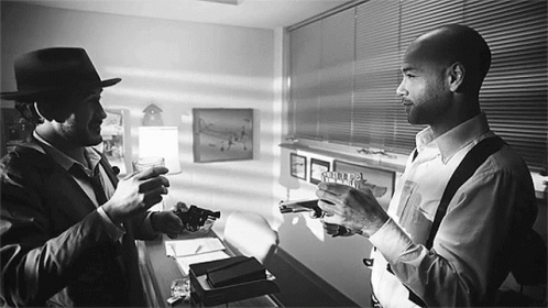 A Man In A Hat Holds A Gun Next To Another Man Who Holds A Gun GIF