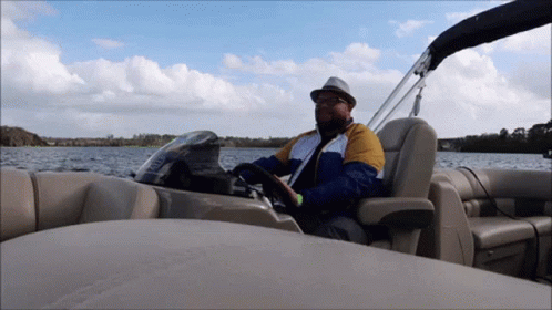 A Man In A Hat Is Driving A Pontoon Boat On A Lake GIF