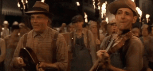 A Man In A Hat Is Playing A Violin In Front Of A Crowd Of People GIF