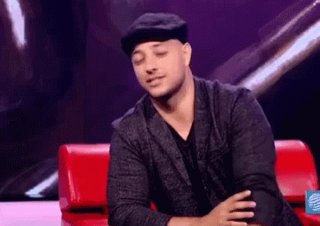 A Man In A Hat Is Sitting On A Red Couch With His Hands Folded . GIF
