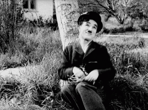A Man In A Hat Is Sitting Under A Tree In The Grass . GIF