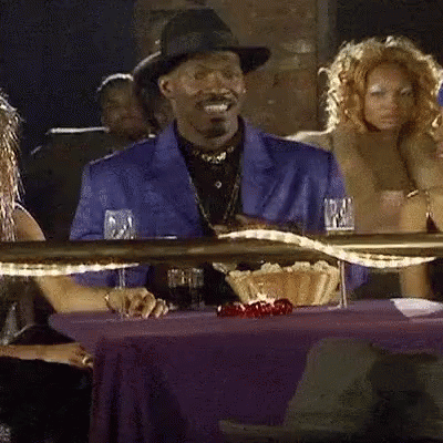 A Man In A Hat Is Smiling While Sitting At A Table With A Purple Table Cloth And Glasses Of Wine GIF