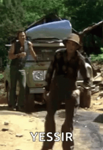 A Man In A Hat Is Standing In Front Of A Car With A Canoe On Top Of It . GIF