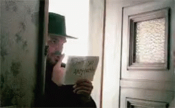 A Man In A Hat Is Standing In Front Of A Door Holding A Piece Of Paper That Says Do N't Say Anything GIF