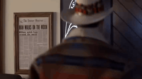 A Man In A Hat Is Standing In Front Of A Framed Newspaper That Says Man Walks On The Moon GIF