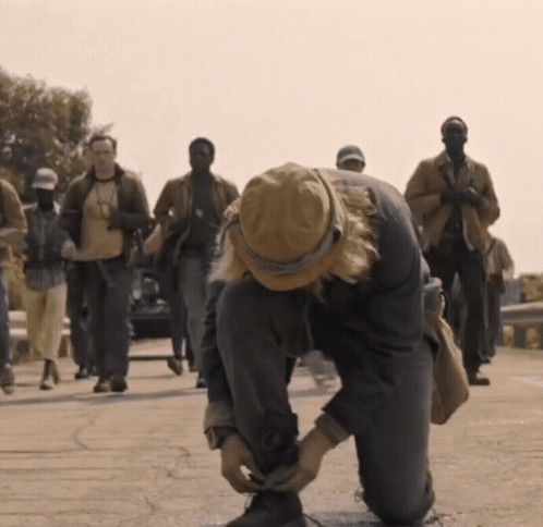 A Man In A Hat Is Tying His Shoelaces While A Group Of People Walk Behind Him GIF